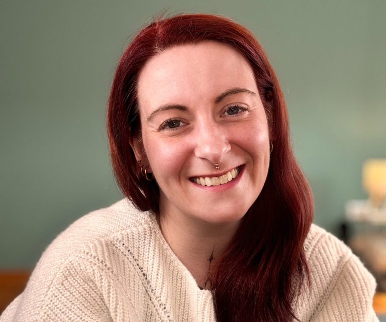 Lächelnde Meike mit rotem Haar, trägt einen hellen Pullover, vor einem grünen Hintergrund.