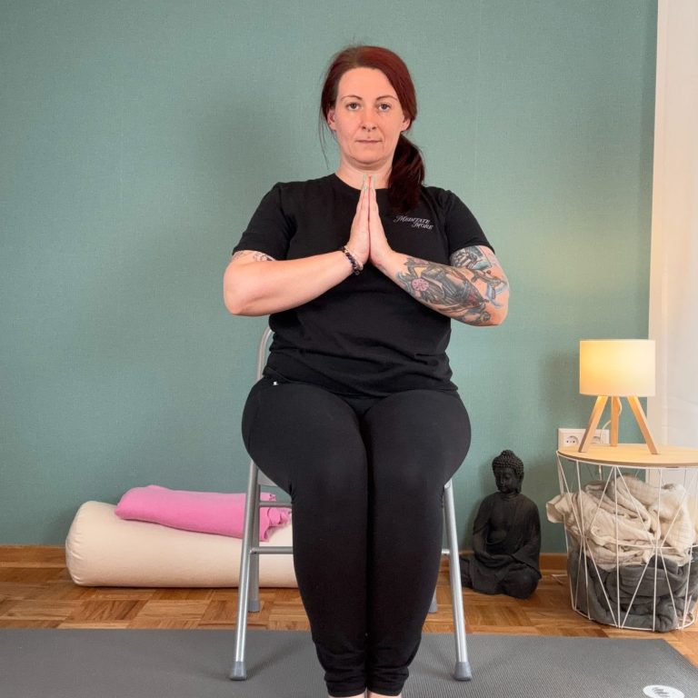 Frau in Schwarz sitzt auf einem Stuhl und macht eine Yoga-Pose mit gefalteten Händen.