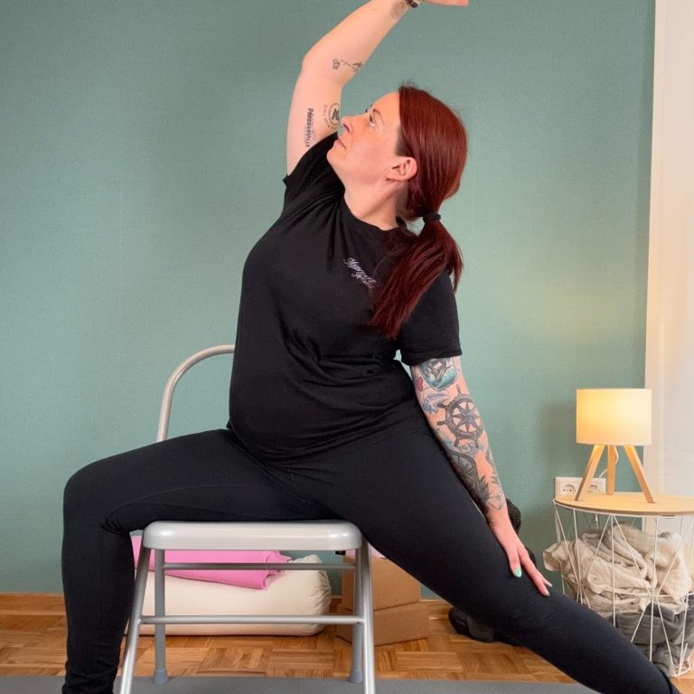 Frau in sportlicher Kleidung übt den Krieger 2 im Sitzen auf einem Stuhl.
